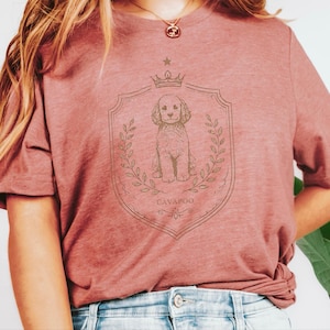 May include: A dusty rose short-sleeved t-shirt featuring a brown line-art graphic of a Cavapoo dog centred within a decorative shield crest, flanked by laurel branches, topped with a small crown, and the text CAVAPOO printed below.