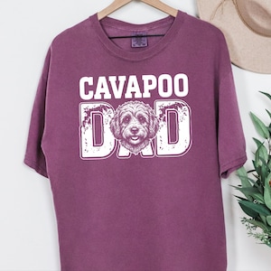 May include: A plum-colored short-sleeve t-shirt featuring the white text CAVAPOO DAD printed on the front. The word DAD incorporates a detailed line-art illustration of a Cavapoo dog face in place of the letter A, set against a distressed background.