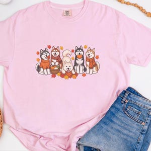 May include: A light pink short-sleeved t-shirt featuring a graphic print of five husky dogs wearing scarves and sweaters, surrounded by autumn leaves and pumpkins. The shirt has a crew neckline and a small brand label at the inner collar.