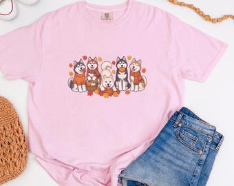 May include: A light pink short-sleeved t-shirt featuring a graphic print of five husky dogs wearing scarves and sweaters, surrounded by autumn leaves and pumpkins. The shirt has a crew neckline and a small brand label at the inner collar.