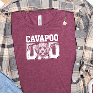 May include: A maroon short-sleeved t-shirt featuring the white text CAVAPOO DAD with a graphic of a dog face replacing the letter A. The shirt is layered under a tan, grey, and black plaid button-down shirt, blue denim, and white trainers.