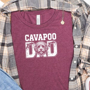 May include: A maroon t-shirt featuring the white text CAVAPOO DAD with a graphic of a dog face replacing the letter A. The shirt is layered under a tan, gray, and black plaid button-down shirt, with blue denim fabric and white sneakers visible.