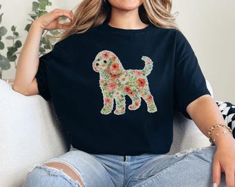 Puede incluir: Una camiseta negra de manga corta con un estampado gráfico de la silueta de un perro rellena de un colorido patrón floral. El diseño incluye varias flores en tonos rosa, rojo, naranja y verde sobre la tela negra sólida de la prenda.