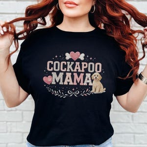 May include: A black short-sleeved t-shirt featuring a graphic print with the text "COCKAPOO MAMA" in pink glittery letters. The design includes a small illustration of a light-colored Cockapoo puppy, pink hearts, a paw print, and a bone.
