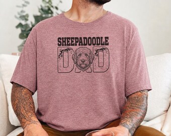 Peut inclure: Un t-shirt à manches courtes mauve chiné avec le texte noir SHEEPADOODLE DAD et un dessin de tête de chien centré dans la lettre A. Le motif présente un aspect vieilli et usé sur le devant du vêtement.