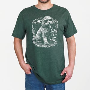 May include: A dark green short-sleeved t-shirt featuring a white graphic print of a curly-haired dog wearing sunglasses and holding a bottle labeled with XXX. The shirt has a crew neckline and a heathered texture throughout the fabric.