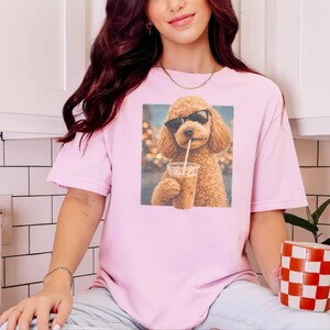 Könnte beinhalten: Ein hellrosa T-Shirt mit kurzen Ärmeln und einem Grafikdruck eines braunen, lockigen Hundes, der eine schwarze Sonnenbrille trägt und einen durchsichtigen Plastikbecher mit einem Strohhalm in den Pfoten hält.