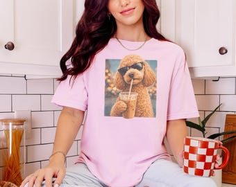 May include: A light pink short-sleeve t-shirt featuring a graphic print of a brown curly-haired dog wearing black sunglasses and holding a clear plastic cup with a straw, set against a blurred background of warm golden lights.