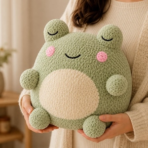 May include: A round, sage green crocheted plush toy shaped like a frog. It features two small ears, a large cream-colored circular belly patch, two small arms, two small legs, and a stitched face with closed eyes, a curved mouth, and pink cheeks.