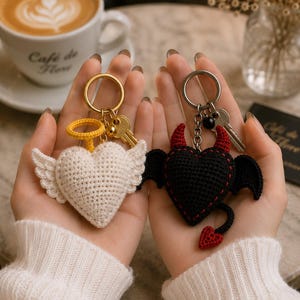 May include: Two crochet heart keychains, one white with wings and a halo, the other black with horns and a tail. Both have gold keyrings and keys. The keychains are held up in front of a blurred background.