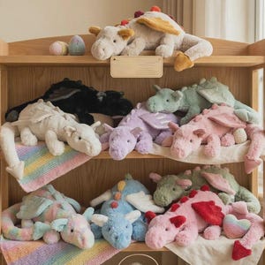 Puede incluir: Una estantería de madera exhibe una colección de juguetes de dragón de peluche en varios colores. Los dragones incluyen blanco, negro, morado, rosa, azul y verde. Algunos dragones tienen alas y están sobre mantas coloridas. También hay huevos decorativos en el estante.