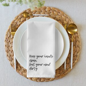 Könnte beinhalten: Eine weiße Stoffserviette mit dem Text "Keep your hands clean, but your mind dirty" in schwarzer Schrift. Die Serviette liegt auf einem weißen Teller, mit goldfarbenem Besteck und einem gewebten Platzset.