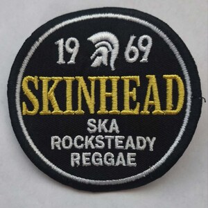 May include: A circular black patch with white and yellow embroidered text. The text reads "SKINHEAD" in yellow, with "1969" and "SKA ROCKSTEADY REGGAE" in white. A white border surrounds the patch.