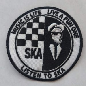 K&ouml;nnte beinhalten: Ein runder, gestickter Aufn&auml;her, der in zwei H&auml;lften geteilt ist. Eine Seite zeigt ein schwarz-wei&szlig;es Schachbrettmuster, die andere eine stilisierte Figur. Der Aufn&auml;her enth&auml;lt das Wort "SKA" und die S&auml;tze "MUSIC IS LIFE", "LIVE A FUN ONE" und "LISTEN TO SKA".