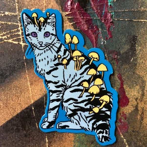 Op de afbeelding: Een blauwe en witte kat sticker met gele paddenstoelen die op zijn rug groeien. De kat heeft paarse ogen en een speelse uitdrukking.