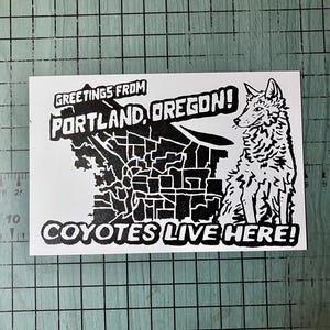 May include: A black and white sticker featuring a stylized illustration of a coyote. The sticker reads "Greetings from Portland, Oregon!" and "Coyotes Live Here!" with a map outline.