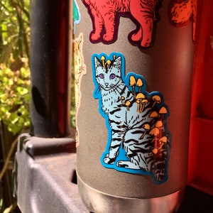 Op de afbeelding: Een grijze metalen waterfles met een blauwe en witte kat sticker. De kat heeft paddenstoelen die op zijn rug groeien.