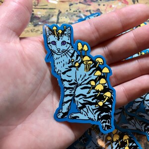 Op de afbeelding: Een blauwe en zwarte kat sticker met gele paddenstoelen die op zijn rug groeien. De sticker is op een blauwe achtergrond.