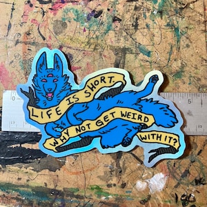 May include: A blue and yellow holographic sticker with a cartoon wolf with three eyes. The wolf is holding a yellow banner that says "Life is short. Why not get weird with it?"