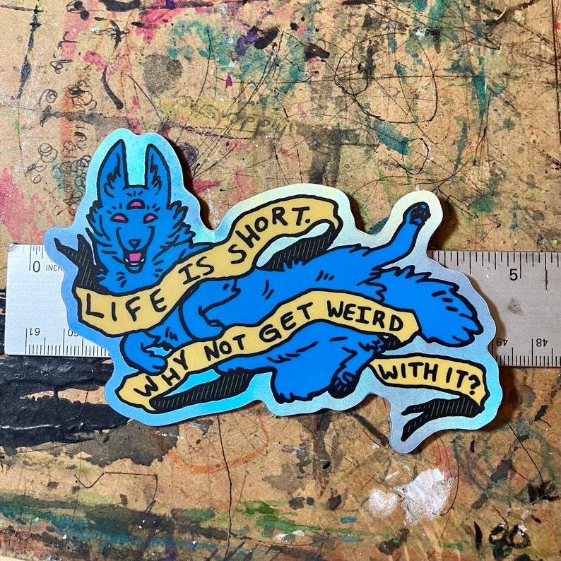 May include: A blue and yellow holographic sticker with a cartoon wolf with three eyes. The wolf is holding a yellow banner that says "Life is short. Why not get weird with it?"