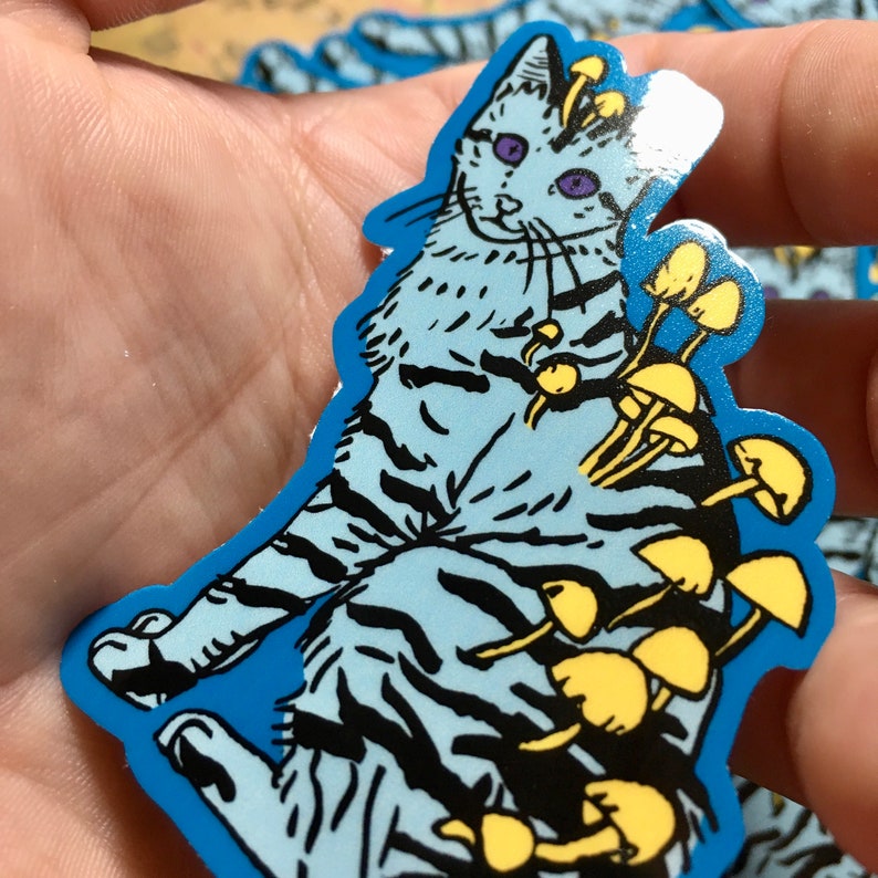 Op de afbeelding: Een blauwe en witte kat sticker met gele paddenstoelen die uit zijn rug groeien. De kat heeft paarse ogen en een zwarte neus.