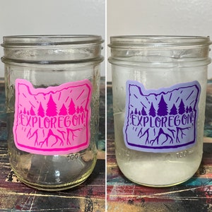 May include: Two glass jars with lids, each with a sticker of the state of Oregon with the text "Explore Oregon" and a silhouette of trees. One sticker is pink and the other is purple.