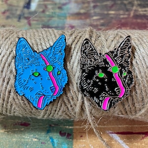 May include: Two enamel pins of a wolf's head with a pink and green stripe down the middle. One wolf is blue and the other is black.