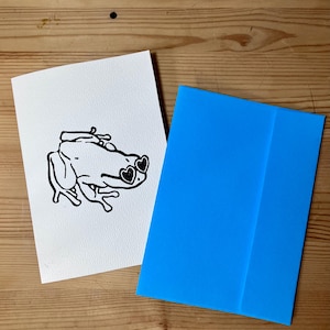 May include: A white card with a black line drawing of a frog wearing heart-shaped sunglasses. Beside it is a bright blue envelope. The frog illustration is on a wooden surface.