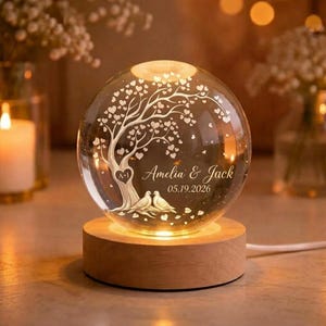 May include: A spherical glass night light on a round wooden base. The glass features an etched design of a tree with heart-shaped leaves, two birds at the base, and the text Amelia & Jack 05.19.2026 with a heart containing the letters A+I.