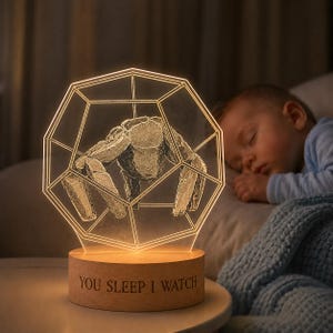 May include: A night light with a geometric design featuring a stone-like creature inside a glowing frame. The light sits on a wooden base with the text "YOU SLEEP I WATCH". The light is illuminating a sleeping baby.
