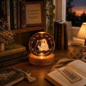 May include: A spherical glass night light featuring an etched illustration of a cat and a ghost reading a book together, surrounded by stars and a crescent moon. The light sits on a round wooden base engraved with the text Just one more chapter.