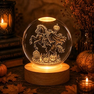 May include: A clear glass sphere lamp featuring an internal 3D laser-etched design of a headless horseman riding a horse, holding a pumpkin, with a tombstone marked RIP, pumpkins, and a fence, set on a round, illuminated wooden base.