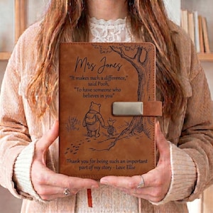 Puede incluir: Un diario de cuero marrón con una ilustración grabada con láser de Winnie the Pooh y Piglet caminando cerca de un árbol. El texto dice: Mrs Jones, It makes such a difference, said Pooh, To have someone who believes in you, Thank you for being such an important part of my story - Love Ellie.