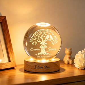 May include: A spherical glass night light resting on a circular wooden base. The glass sphere features an etched design of a tree with two birds, the names Emma and James, and the date EST. 06.25.1976. The wooden base is engraved with I Love You.