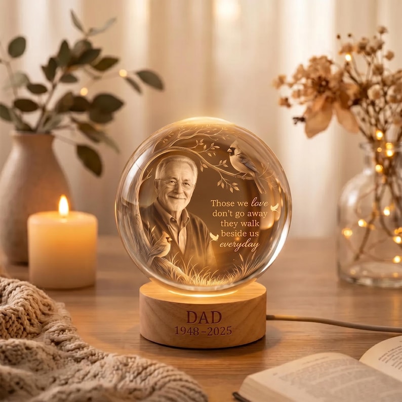 May include: A spherical glass night light featuring an engraved portrait of an older man, a cardinal bird on a branch, and the text: Those we love don't go away they walk beside us everyday. The base is wood with DAD 1948-2025 engraved on it.