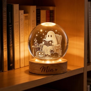 May include: A spherical glass night light featuring an internal laser-etched illustration of a ghost reading a book in an armchair with a cat and a pumpkin. The light sits on a round wooden base engraved with the name Mia and a small heart symbol.