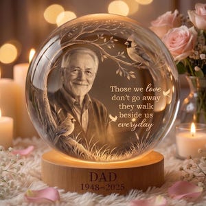 May include: A spherical glass memorial ornament on a wooden base engraved with DAD 1948-2025. The glass features an etched portrait of an older man, two cardinals on tree branches, and the text: Those we love don't go away they walk beside us everyday.