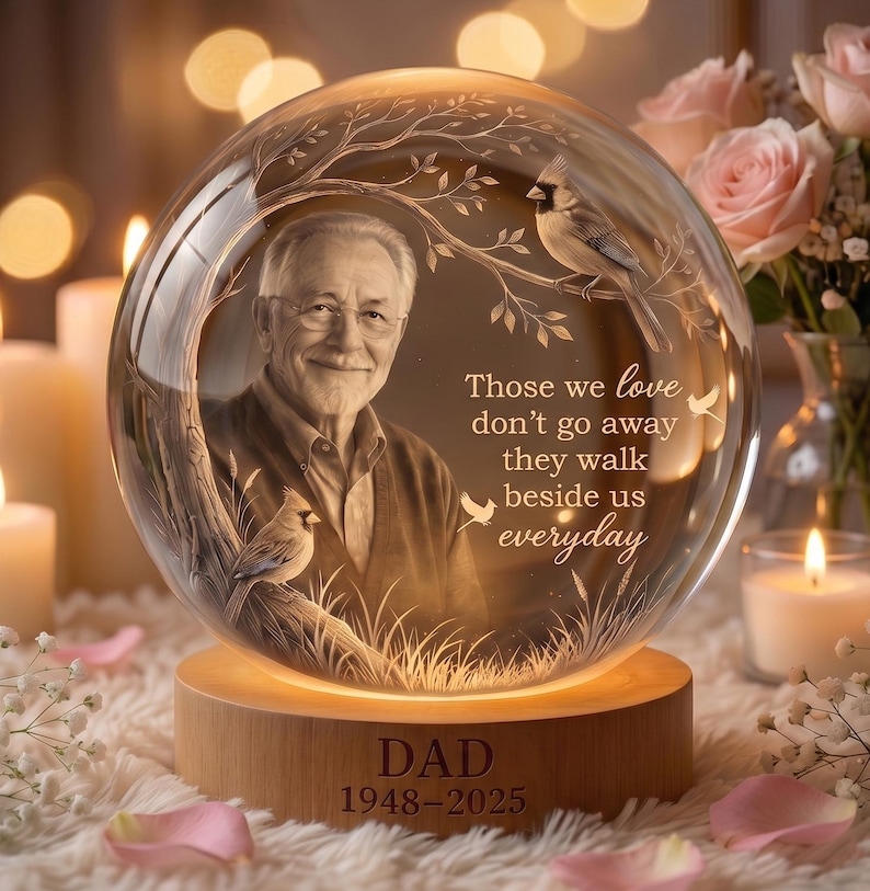 May include: A spherical glass memorial ornament on a wooden base engraved with DAD 1948-2025. The glass features an etched portrait of an older man, two cardinals on tree branches, and the text: Those we love don't go away they walk beside us everyday.
