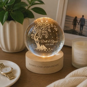 May include: A clear glass sphere lamp resting on a cylindrical wooden base. The sphere features an etched design of a tree with heart-shaped leaves, two birds, the text "Brett & Alyssa" and "6.24.2026". The wooden base is engraved "HAPPY 15TH ANNIVERSARY".
