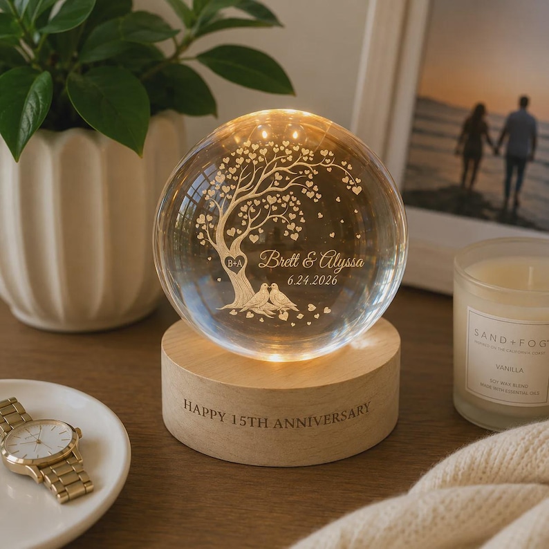 May include: A clear glass sphere lamp resting on a cylindrical wooden base. The sphere features an etched design of a tree with heart-shaped leaves, two birds, the text "Brett & Alyssa" and "6.24.2026". The wooden base is engraved "HAPPY 15TH ANNIVERSARY".