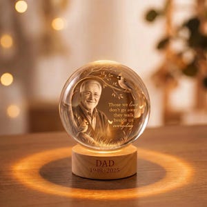May include: A clear glass sphere featuring an etched portrait of an older person, a cardinal bird, and the text: Those we love don't go away they walk beside us everyday. The sphere sits on a light wood base engraved with DAD 1948-2025.