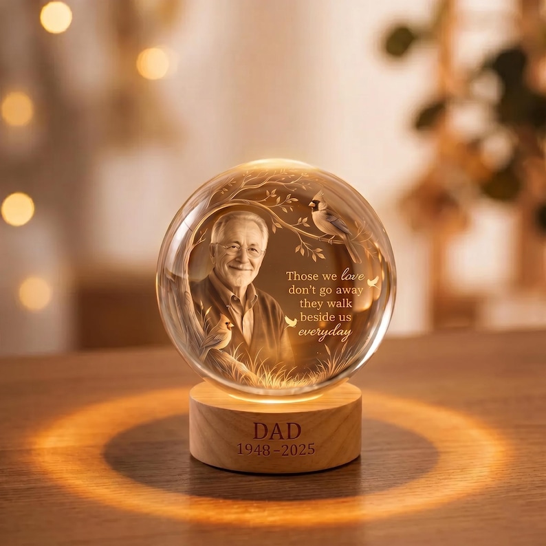 May include: A clear glass sphere featuring an etched portrait of an older person, a cardinal bird, and the text: Those we love don't go away they walk beside us everyday. The sphere sits on a light wood base engraved with DAD 1948-2025.