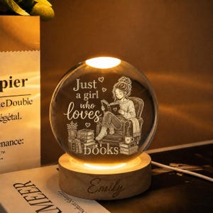 May include: A spherical glass night light on a circular wooden base. The glass features an etched illustration of a person reading in a chair with stacks of books and the text "Just a girl who loves books". The wooden base is engraved with the name "Emily".