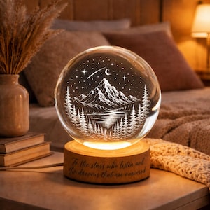 Könnte beinhalten: Ein kugelförmiges Nachtlicht aus Glas mit einer geätzten Berglandschaft, einem See, Tannen und einem Sternenhimmel mit einer Mondsichel. Die Glaskugel ruht auf einem runden Holzsockel, der mit dem Text, To the stars who listen and the dreams that are answered, graviert ist.