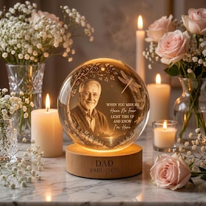 May include: A memorial sphere with a photo of a man, text that reads "WHEN YOU MISS ME Have No Fear LIGHT THIS UP AND KNOW I'm Here", and the word "DAD" with the years 1948-2025. The sphere sits on a wooden base and is surrounded by candles and flowers.