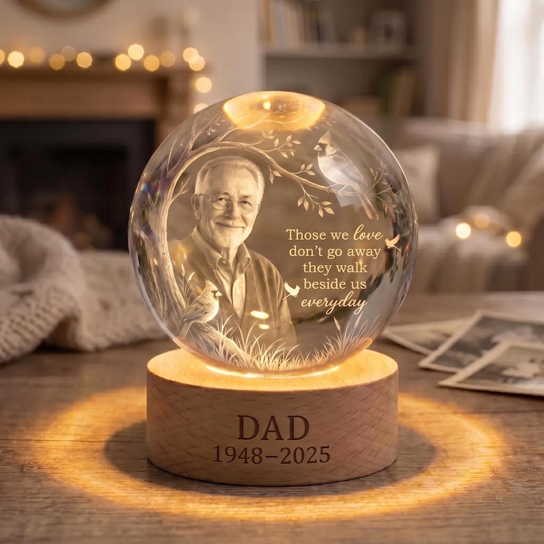 May include: A clear glass sphere featuring an etched portrait of an older man, a cardinal bird on a branch, and the text: Those we love don't go away they walk beside us everyday. The sphere sits on a round wooden base engraved with DAD 1948-2025.