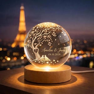 May include: A clear glass sphere lamp on a round wooden base, featuring an etched design of a tree with heart-shaped leaves, two birds at the base, and the text Amelia & James 04.16.2026 with a heart containing the initials A+I on the tree trunk.