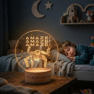 Może przedstawiać: Dekoracyjna lampka nocna ze złotym wzorem owada i słowem "AMAZE!" powtórzonym trzy razy. Podstawa lampki jest drewniana i ma napis "YOU SLEEP I WATCH". Światło znajduje się na drewnianym stole w pokoju dziecięcym.