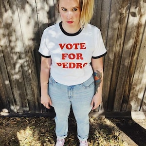 Op de afbeelding: Wit T-shirt met zwarte rand met de rode tekst "VOTE FOR PEDRO". De persoon draagt een lichtblauwe spijkerbroek en roze sneakers. De achtergrond is een houten hek.