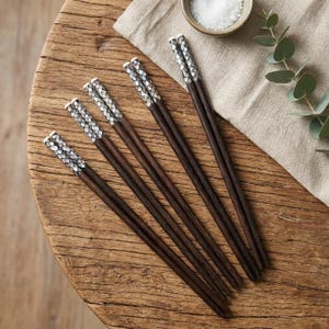 Op de afbeelding: Een set van vijf donkerbruine houten eetstokjes met decoratieve zilver-witte patronen aan de uiteinden. De eetstokjes liggen op een rustieke houten tafel, met een klein bakje zout en groen in de achtergrond. De eetstokjes zijn ongeveer 23 cm lang.